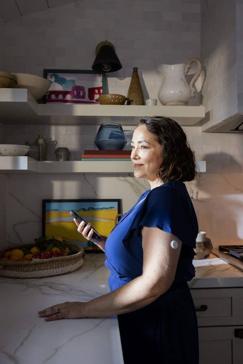 a commercial photograph of a woman standing in her kitchen wearing her Abbott CGM Sensor